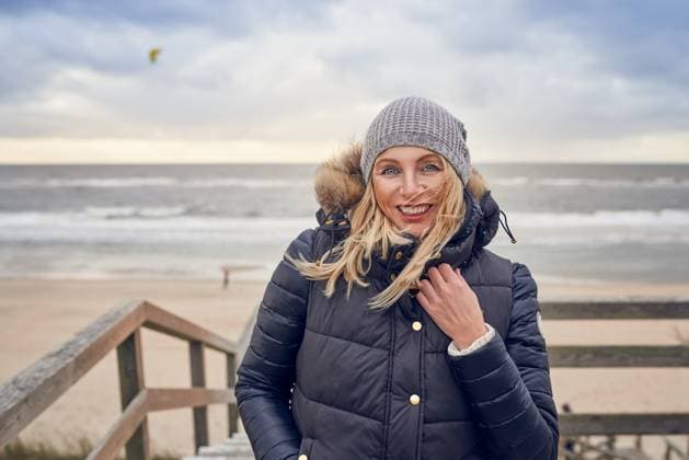 Middle-aged woman braving a cold winter day at the sea Middle-aged woman braving a cold winter day at the seaside standing on a wooden deck overlooking the beach on a breezy day smiling happily at the camera Copyright: xZoonar.com LarsxZahnerx 11589711 ,model released, Symbolfoto ,property released