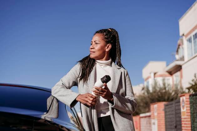 Thoughtful woman standing by car and holding electric plug at vehicle charging station model released, Symbolfoto, EBBF07931