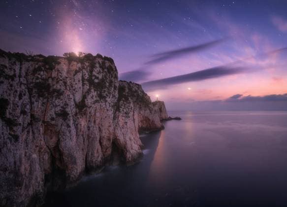 RECORD DATE NOT STATED Lighthouse on the mountain peak at starry night in summer. Beautiful cliffs, rocky sea coast, bright stars, milky way and violet sky with clouds at dusk. Lighthouse of Cape Lefkada, Greece. Landscape