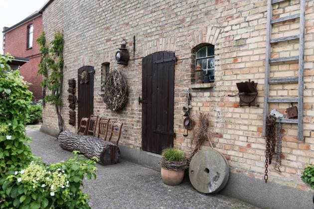 Stadtgarten in Neukloster in Mecklenburg-Vorpommern Scheune aus Ziegelsteinen mit zwei Holztüren und einer Holzleiter sowie alten Ofentueren an der Wand., davor aus einem Baumstamm und Holzlehnen von Stuehlen gebaute Bank. - Ein Garten versteckt hinter einem Backsteinhaus in der Kleinstadt Neukloster direkt am Hopfenbach. Stauden, Gehoelze, Rosen, Obstbäume und Gemuesebeete sind dekoriert mit altem Trödel. An verschiedenen Ecken laden Sitzgelegenheiten zum Entspannen ein. *** City Garden in Neukloster in Mecklenburg Western Pomerania Barn from Bricks with Two Wooden doors and a Wood ladder and Old oven doors to the Wall , before that from a Tree trunk and Wood backs from Chairs built Bank a Garden hidden behind a Brick house in t
