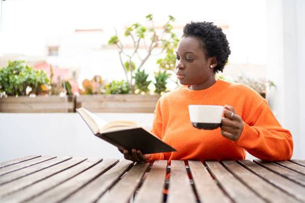 IMAGO Creative, Lifestyle, Tik Tok-Trend thatgirl Young African American woman in orange sweater sitting at table with cup of hot drink and reading book OscarDeDios_ID13083_398484_009 Copyright: xOscarxDexDiosx Model Released