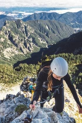 Vertical photo of woman mountaineer with white helmet looking down, climbing on via ferrata trail in the Alps Caucasian woman mountaineer climbing up the mountain on via ferrata trail, high up in the mountain peaks. Woman wearing a protective head gear and uses a safety harness while she climbs. ,model released, Symbolfoto Copyright: xZoonar.com ZigaxPlahutarx 19685814 ,model released, Symbolfoto ,property released