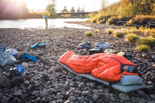 Woman asleep in sleeping bag on river edge with fiisherman behind Shelburne, New Hampshire, United States CR_BRDO2302087A-1123611-01 ,model released, Symbolfoto
