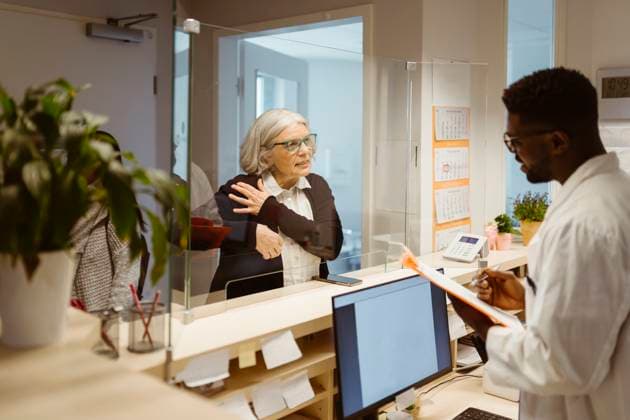 Senior female patient explaining health issues to male receptionist through transparent shield in clinic Germany, Berlin, model released, property released Copyright: xMaskotx , MA101287 MA101287