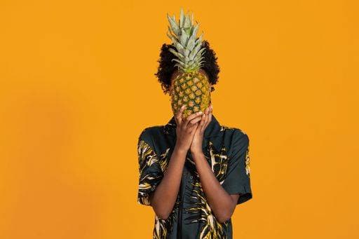 A Young African American woman with short hair dressed in a summer shirt posing with pineapple on an orange colored background *** einer Jung afrikanisch amerikanisch Frau mit Kurze Haare angezogen ÊIn einer Sommer Hemd posierend mit Ananas am An&Verkauf Orangefarbene farbige Hintergrund 8442344 ,model released, Symbolfoto RECORD DATE NOT STATED