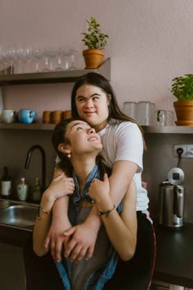 Young woman with down syndrome hugging sister from behind in kitchen at home Germany, Berlin, model released, property released Copyright: xMaskotx , MA102442 MA102442