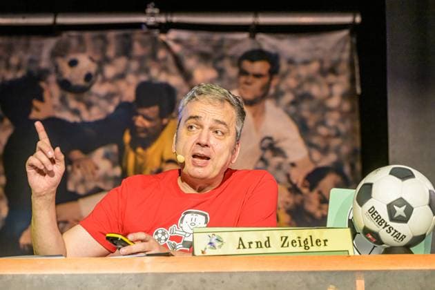 Zeiglers wunderbare Welt des Fußballs 27.09.2023, Uni Aula, Vechta, Zeiglers wunderbare Welt des Fußballs, Der Moderator strickt Fußballträume: Witzig, traurig, skurril und immer auch ein bisschen böse. Offen und unverblümt redet er in seinem Arbeitszimmer über Tragik, Tränen und Triumphe im Bild Arnd Zeigler (Moderator, ) an seinem Bühnenschreibtisch in der Aula der Uni Vechta vor ausverkauften Haus.