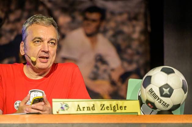 Zeiglers wunderbare Welt des Fußballs 27.09.2023, Uni Aula, Vechta, Zeiglers wunderbare Welt des Fußballs, Der Moderator strickt Fußballträume: Witzig, traurig, skurril und immer auch ein bisschen böse. Offen und unverblümt redet er in seinem Arbeitszimmer über Tragik, Tränen und Triumphe im Bild Arnd Zeigler (Moderator, ) an seinem Bühnenschreibtisch in der Aula der Uni Vechta vor ausverkauften Haus.