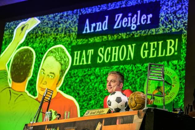 Zeiglers wunderbare Welt des Fußballs 27.09.2023, Uni Aula, Vechta, Zeiglers wunderbare Welt des Fußballs, Der Moderator strickt Fußballträume: Witzig, traurig, skurril und immer auch ein bisschen böse. Offen und unverblümt redet er in seinem Arbeitszimmer über Tragik, Tränen und Triumphe im Bild Arnd Zeigler (Moderator, ) an seinem Bühnenschreibtisch in der Aula der Uni Vechta vor ausverkauften Haus.