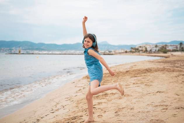 Cheerful girl wearing wireless headphones listening to music and dancing on coastline at beach model released, Symbolfoto, JOSEF20096