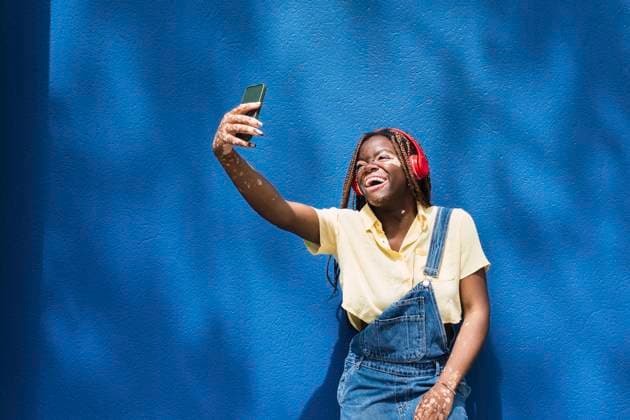 Happy young woman taking selfie through smart phone in front of blue wall model released, Symbolfoto, PNAF06061