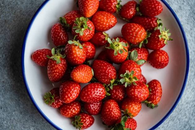 Top view of bright red organic strawberries with green leaves, displayed in a white plate with a blue rim, against a textured gray backdrop. Model Released AnnaBogush_ID17230_524310_003 Copyright: xAnnaxBogushx RECORD DATE NOT STATED