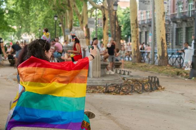 IMAGO Creative: Sommer, Sommertage im Stadtpark MR_ID17208_15471_JimenaPardo.jpg Side view of woman wraps herself with a large and colorful LGBTQ pride flag while taking selfie with her smartphone in a park with people in the background Model Released NataliaPenado_ID17208_523683_015 Copyright: xNataliaxPenadox RECORD DATE NOT STATED