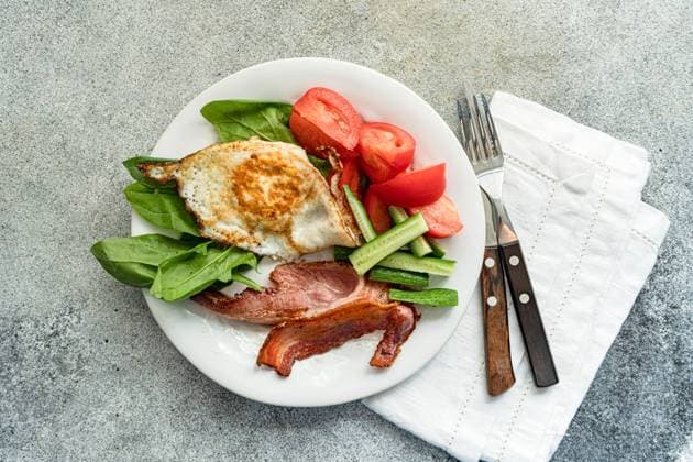 From above of balanced breakfast arrangement on a white plate with fried egg, crispy bacon, fresh spinach leaves, sliced cucumber and wedged tomatoes served with cutlery on grey textured surface Model Released AnnaBogush_ID17558_531810_002 Copyright: xAnnaxBogushx RECORD DATE NOT STATED