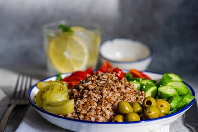 Closeup of healthy lunch bowl with boiled organic buckwheat, fresh cucumber and tomato, and fermented tomato and olives served with glass of pure water with lemon, ice and mint against blurred gray surface Model Released AnnaBogush_ID17559_531818_002 Copyright: xAnnaxBogushx RECORD DATE NOT STATED