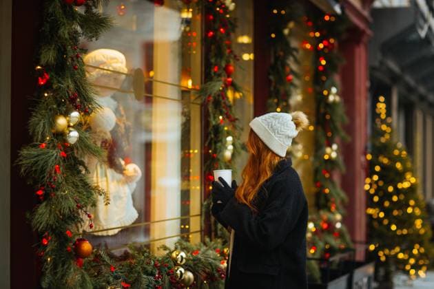 A young woman in a white knit hat and black coat holds a coffee mug while gazing at a festive Christmas window display adorned with lights and decorations in Quebec, Canada Model Released CamilleGirard_ID13407_408511_026 Copyright: xCamillexGirardx