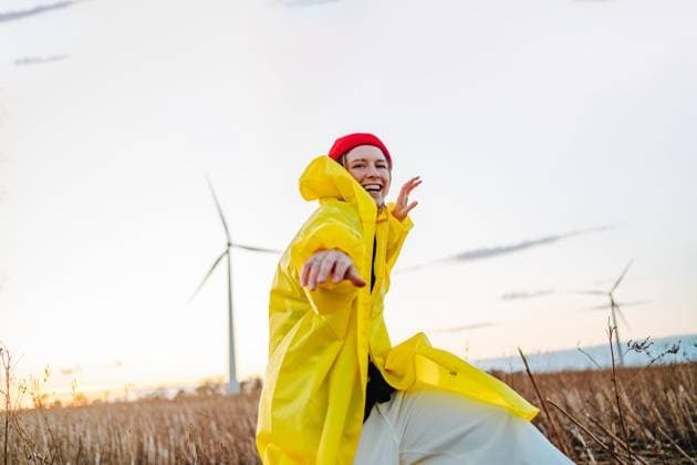 Creative Highlights Symbolbilder Joyful young woman in a vibrant yellow raincoat and red beanie is dancing in a field during sunset with wind turbines in the background. Model Released CaramelFilms_ID17449_529723_019 Copyright: xCaramelxFilmsx RECORD DATE NOT STATED