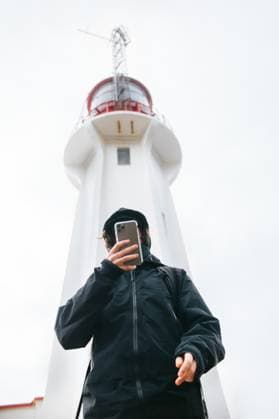 Creative Highlights Symbolbilder A person stands in front of a towering lighthouse on Vancouver Island, engaged with their smartphone, possibly capturing the scene or navigating. Model Released CamilleGirard_ID17696_535225_004 Copyright: xCamillexGirardx