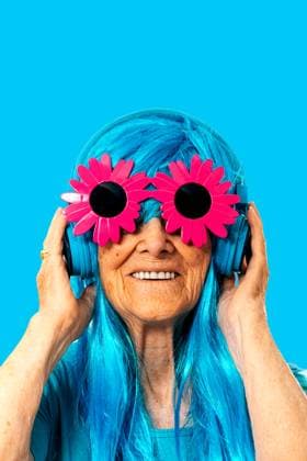 Creative Highlights Symbolbilder Portrait of happy elderly female with wrinkles and toothy smile looking at camera while standing in blue wig with headphones and wearing flower frame sunglasses against blue background in light Model Released SergioVictorVega_ID17016_518843_021 Copyright: xSergioxVictorxVegax
