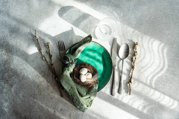 Top view of Easter table setting, showcasing a vibrant green ceramic plate with a small nest with speckled eggs and feathers, placed on gray surface with napkin, cutlery and glass of water Model Released AnnaBogush_ID18583_553999_006 Copyright: xAnnaxBogushx