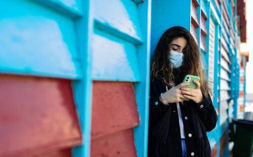 A Caucasian woman with protective mask browsing her phone while standing against a blue metal door 1929210 ,model released, Symbolfoto RECORD DATE NOT STATED