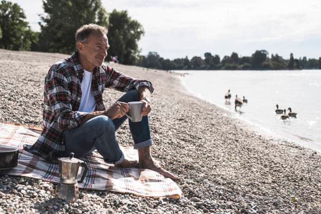 IMAGO Creative, Ostern, Bräuche, Osterspaziergang Thoughtful senior man with coffee cup sitting on blanket at beach model released, Symbolfoto, UUF30848