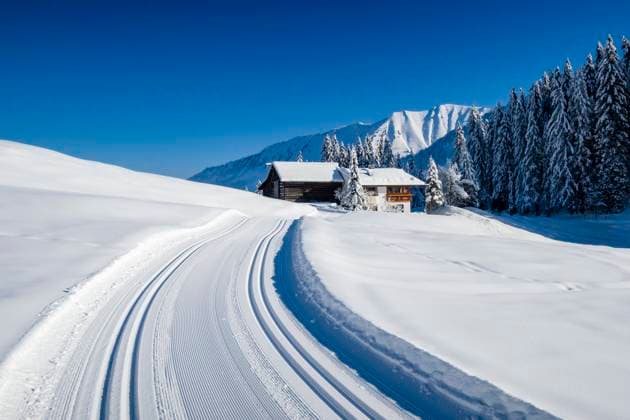 A freshly groomed cross-country ski trail in a winter landscape in Kleinwalsertal valley, a farmhouse and snow-covered mountains in the distance. Mittelberg Vorarlberg Austria FB_2024_0003