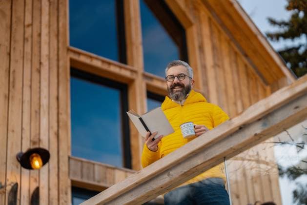 Man standing on cabins patio, drinking hot tea, coffee and reading book. Handsome man spending relaxing, stress-free winter weekend in cabin in mountains, enjoying alone time. Mature man standing on cabins patio, drinking hot tea, coffee and reading book. Handsome man spending relaxing, stress-free winter weekend in cabin in mountains, enjoying alone time. model released