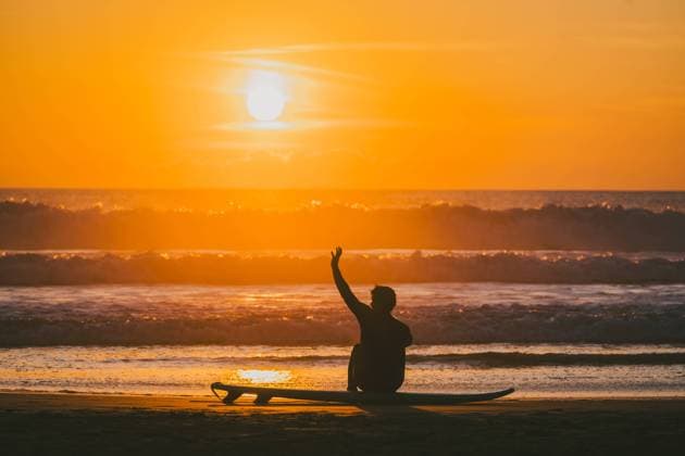 Unrecognizable male surfer sitting with raised hand on surfboard by sea at sunset Silhouette of unrecognizable male surfer sitting on surfboard with raised arm while enjoying picturesque sunset view over waving sea at evening Model Released AlvaroMontero_ID19256_573102_004 Copyright: xAlvaroxMonterox