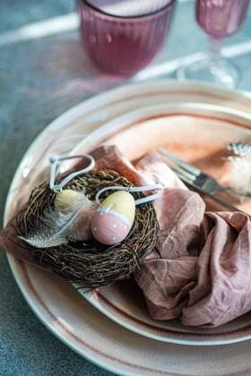 An artistic Easter table setting featuring a nest with pastel decorated eggs atop ceramic plates, complemented by a dusty pink napkin. Model Released AnnaBogush_ID19406_575233_002 Copyright: xAnnaxBogushx