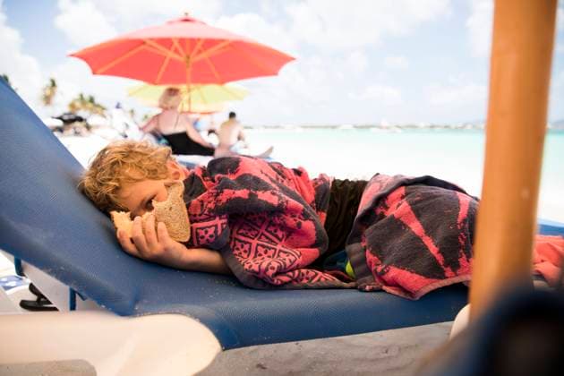 Boy Holding Sandwich sits on Lounge Chair Under Beach Umbrella Blowing Point Village, Anguilla CR_EMJA240225-109049-01 ,model released, Symbolfoto