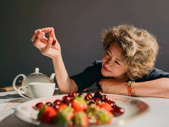 Creative Highlights Symbolbilder Smiling mature woman picking up cherry from plate on table at home model released, Symbolfoto, IEF00535