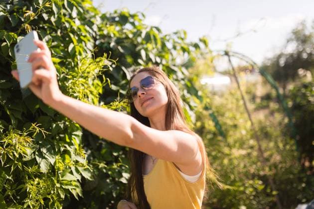 IMAGO Creative: Sommer, Sommertage im Garten MR_ID19530_16353_MarinaGabitova.jpg A cheerful young woman in a yellow tank top takes a selfie with her phone among lush greenery on a sunny day Model Released Declaria_ID19530_579187_001 Copyright: xDeclariax RECORD DATE NOT STATED
