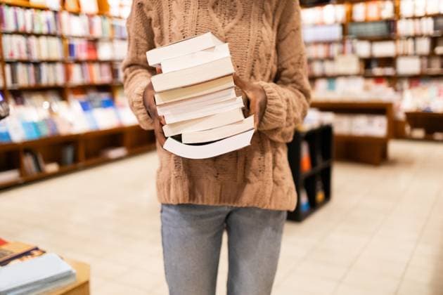 Close-up image of an unrecognizable individual in a cozy sweater holding a large pile of books, standing in the aisle of a well-stocked library Model Released DavidMunoz_ID20111_595378_042 Copyright: xDavidxMunozx