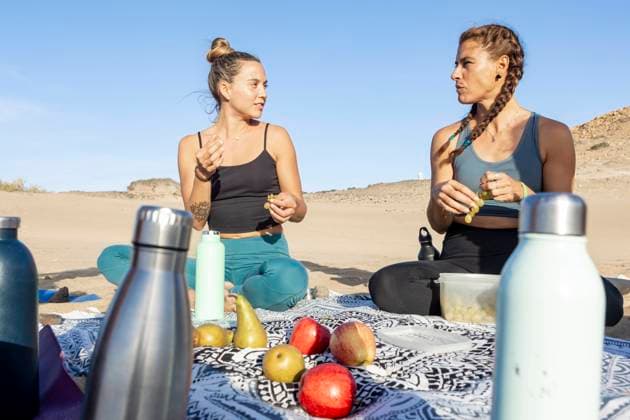 Two women enjoying a nutritious picnic on a beach mat, surrounded by fresh fruit and water bottles, engaged in a conversation with a clear blue sky above. Model Released JuanAizpuru_ID19654_582744_024 Copyright: xJuanxAizpurux