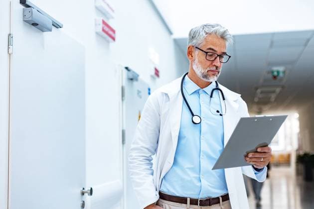 Portrait of doctor in hospital corridor. Handsome doctor with gray hair wearing white coat, stethoscope around neck standing in modern private clinic. Portrait of mature doctor in hospital corridor. Handsome doctor with gray hair wearing white coat, stethoscope around neck standing in modern private clinic. model released