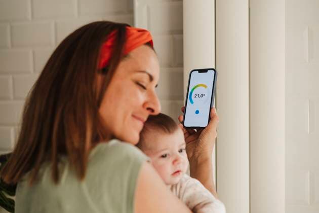 IMAGO Creative, Jahreszeit Sommer, Hitze Mother embracing baby daughter and adjusting radiator through smart phone at home model released, Symbolfoto property released, EBBF08730