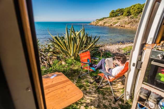 Blick aus der Schiebetüre eines Wohnmobils das auf auf einem Stellplatz am unbefestigtem Ufer an der Mittelmeerküste steht, eine junge Frau sitzt auf Campingstühlen und liest in einem Buch. Sizilien, Italien. Ein Wohnmobil steht auf einem Stellplatz am Ufer des Mittelmeers. *** View from the sliding door of a motorhome standing on a pitch on the unpaved shore on the Mediterranean coast, a young woman sits on camping chairs and reads a book Sicily, Italy A motorhome stands on a pitch on the shore of the Mediterranean Sea