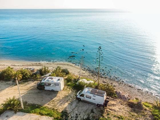 Ein Campingbus und ein Alkoven Wohnmobil stehen auf einem Stellplatz am unbefestigtem Ufer an der Mittelmeerküste, Boote liegen davor am Strand. Sizilien, Italien. Ein Wohnmobil steht auf einem Stellplatz am Ufer des Mittelmeers. *** A camper van and an alcove motorhome are parked on a pitch on the unpaved shore on the Mediterranean coast, boats lie in front of them on the beach Sicily, Italy A motorhome is parked on a pitch on the shore of the Mediterranean Sea