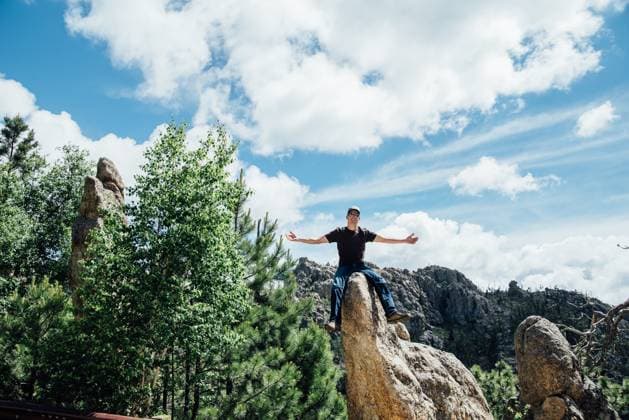 Man with arms outstretched sitting on rocks in sunny mountain scene Keystone, South Dakota, United States CR_KIFR240402-1377296-01 ,model released, Symbolfoto