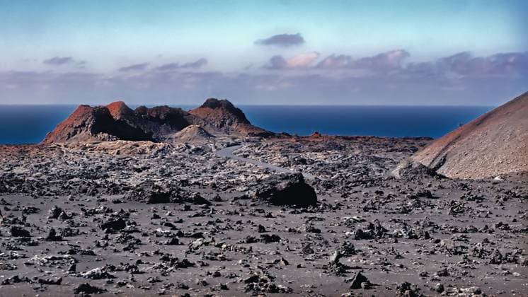 Moon-like volcanic landscape in the heart of the fire mountains of Lanzarote A moon-like, unreal landscape in the heart of the volcanic island of Lanzarote, in the Fire Mountains or Timanfaya Mountains. Black lava stones, a crater opening of a small volcanic cone in the evening light. Behind the dark blue Atlantic and a few gray-purple clouds. Copyright: xZoonar.com AkimaxFuturax 20691995