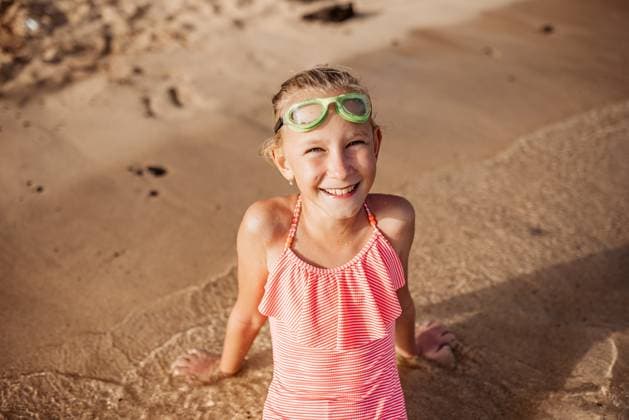 Young girl playing, swiming and splashing in fresh sea water. Smilling blonde girl in swimsuit with and swimming goggles. Concept of beach summer vacation with kids. Young girl on the beach. Smilling blonde girl enjoying sandy beach, looking at crystalline sea in Canary Islands. Concept of beach summer vacation with kids. model released