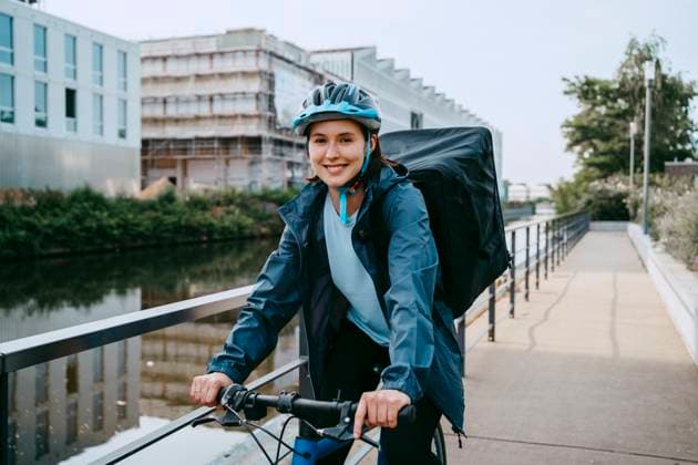 Portrait of smiling female food delivery person cycling on footpath near canal in city Germany, Berlin, model released Copyright: xMaskotx , MA108243 MA108243