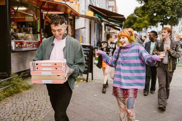Creative Highlights Symbolbilder Smiling non-binary person holding stack of pizza boxes while walking with friends at street in city Germany, Berlin, model released Copyright: xMaskotx , MA108289 MA108289