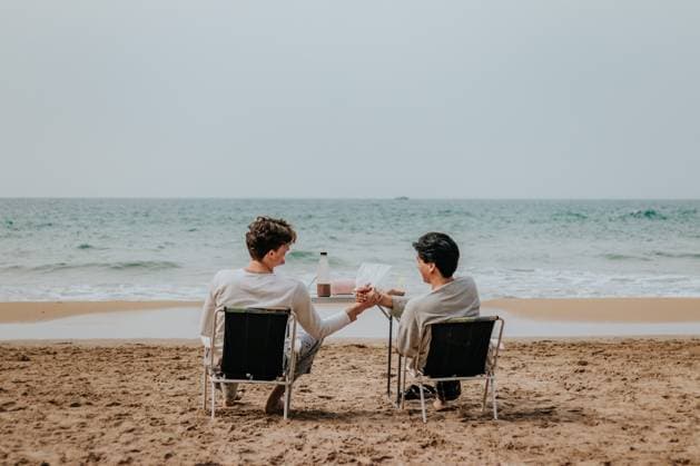 Creative Highlights Symbolbilder MR_ID20863_16993_SergioPauloHerreraMorales.jpg, MR_ID20863_16994_JackRudy.jpg Back view of two friends toast drinks while seated at a table on a sandy beach, with the ocean in the background. Model Released DavidGari_ID20863_615444_004 Copyright: xDavidxGarix