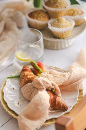 A cozy Easter brunch setting featuring freshly baked croissants, coconut muffins on a ceramic plate, with spring-inspired tableware and a hint of spring flowers, creating a warm, inviting atmosphere for friends and family Model Released JoannaFufal_ID20588_608225_004 Copyright: xJoannaxFufalx