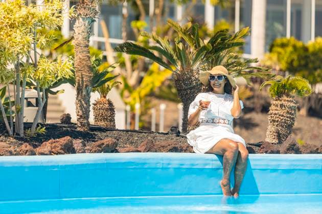 A smiling middle aged hispanic woman in a white dress and sunhat sits at the edge of a pool, holding a cocktail, surrounded by exotic plants. Model Released JuanAizpuru_ID20788_613331_028 Copyright: xJuanxAizpurux