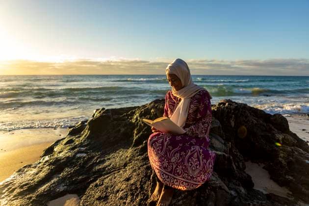 Creative Highlights Symbolbilder A peaceful scene of a Muslim woman engrossed in reading a book, seated on the rocks by the beach with waves and the sunset in the background Model Released JuanAizpuru_ID20807_613625_028 Copyright: xJuanxAizpurux