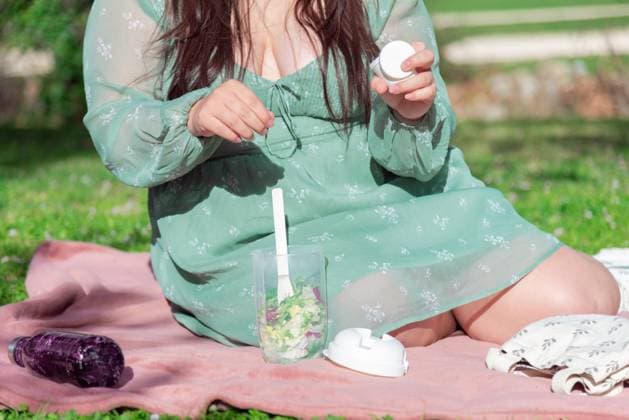 A woman in a green dress is seated on a pink blanket outside, preparing to eat a fresh salad from a clear container, with a sunny backdrop. Model Released NataliaPenado_ID20535_606443_014 Copyright: xNataliaxPenadox RECORD DATE NOT STATED