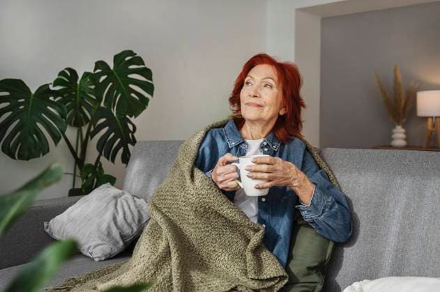 Senior woman sitting relaxed under blanket on sofa drinking tea at home model released, Symbolfoto property released, ALKF01057